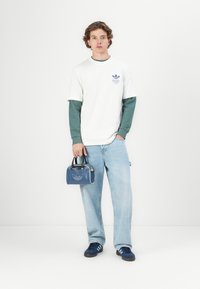 Jeune homme portant des chemises à manches longues superposées blanches et vertes, un jean bleu clair, des baskets marines, tenant un petit sac à main bleu avec un logo sur fond blanc.