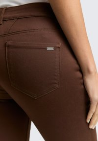 Bruine spijkerbroek van denim met een enkele achterzak met een metalen logo-accent. De gladde textuur en de aansluitende pasvorm verbeteren de algehele uitstraling.