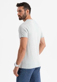 Světle šedé tričko s krátkým rukávem a kulatým výstřihem, vyrobené z měkké tkaniny, v kombinaci s modrými džínami. Jednoduchý, nenápadný design.