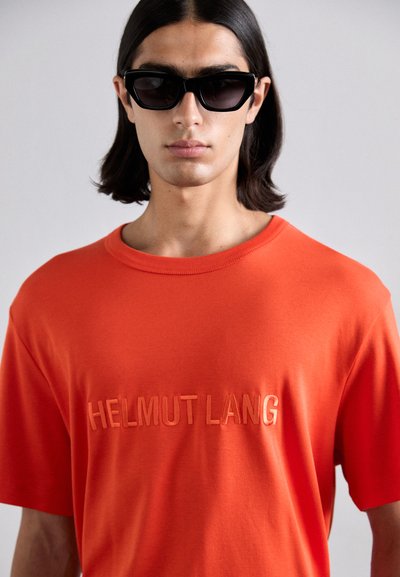 Camiseta de algodón naranja con un logo en relieve, cuello redondo y mangas cortas. Llevada con gafas de sol negras de forma angular.