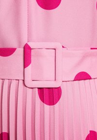 Robe rose ornée de grands pois fuchsia, avec une jupe plissée et une boucle de ceinture carrée assortie, fabriquée en tissu léger.