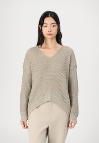 Maglione lavorato a maglia color beige chiaro con scollo a V, spalle cadute e orlo asimmetrico. Tessuto texturizzato con una vestibilità ampia. Abbinato a pantaloni beige.