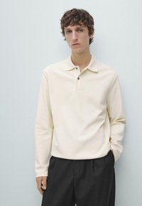 Koszulka polo z długim rękawem wykonana z miękkiej kremowej tkaniny, z kołnierzykiem, plisą na trzy guziki i luźnym krojem, zestawiona z ciemnymi, eleganckimi spodniami.