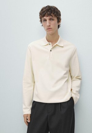 Polo a maniche lunghe in morbido tessuto color crema, con colletto, abbottonatura a tre bottoni e vestibilità rilassata, abbinata a pantaloni scuri sartoriali.