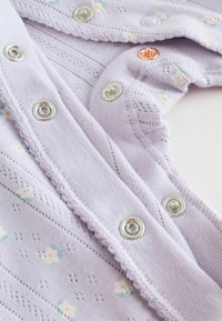 Lila bomullsbody med blommönster, silvriga tryckknappar och en vågig kant längs halsringningen. Mjuk textur med en lätt stickad design.