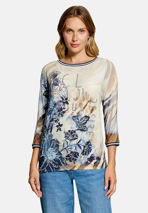 Frau trägt langärmeliges beiges Oberteil mit blauem Blumenmuster und Schriftzug, kombiniert mit blauen Jeans, steht vor weißem Hintergrund.
