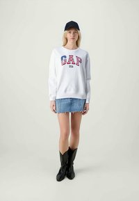 Biely sveter s grafiku "GAP USA" v červenej a modrej farbe, skombinovaný s džínsovou mini sukňou a čiernymi vysokými čižmami. Čierna čiapka na hlave.