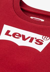 Czerwona bluza z dużym białym logo Levi's na przodzie. Materiał wydaje się miękki, z prążkowanym dekoltem dla większego komfortu.