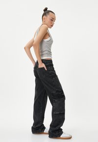 Zwarte spijkerbroek cargo met zijzakken, een relaxte pasvorm en gestructureerde stof. Gekleed met een lichtgrijze tanktop en witte sneakers.