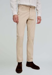 Pantalones de algodón beige con corte ajustado, que presentan un cierre de botón y bolsillos laterales, combinados con botas de ante marrón oscuro hasta el tobillo.