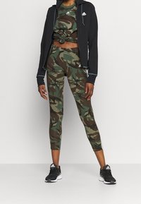 Sada sportovního oblečení Adidas s černou zipovou mikinou, kamuflážním crop topem a legínami. Obsahuje kamuflážový vzor v zelených a hnědých odstínech, doplněno černými teniskami.