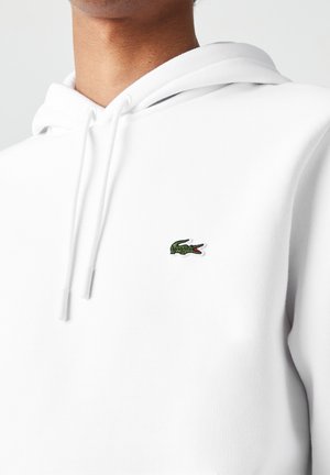 Gros plan d'une personne portant un sweat à capuche blanc avec un petit logo de crocodile vert brodé sur la poitrine.