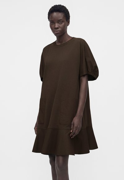 Γυναίκα που φοράει ένα φαρδύ σκούρο καφέ φόρεμα με φουσκωτά κοντά μανίκια, μπροστινές τσέπες και βολάν στο τελείωμα, στέκεται μπροστά σε απλό φόντο.