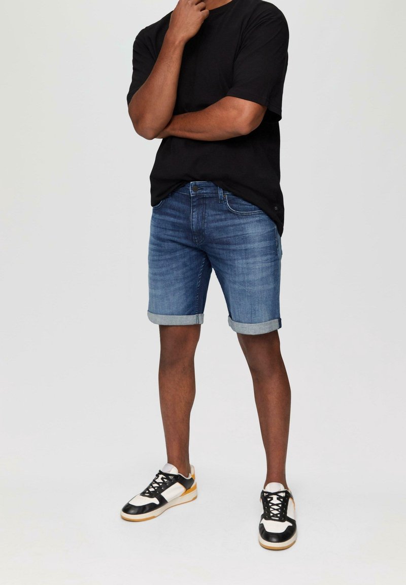 Shorts di denim blu chiaro con risvolti, abbinati a una maglietta nera e scarpe da ginnastica nere con dettagli bianchi e gialli.