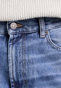 Ljusblå denimjeans med rak skärning, med kontrasterande sömmar, metallknappstängning och en liten logotyp-tagg nära midjan.