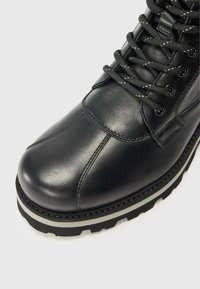 Bota de cuero negro con puntera redondeada, diseño de cordones y suela resistente, ubicada sobre un fondo blanco liso.
