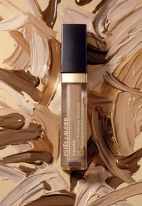 Estee Lauder Futurist concealer i en genomskinlig tub med ett svart fyrkantigt lock, mot bakgrund av virvlande beige och bruna krämtexturer.