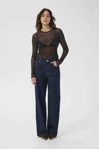 Vrouw staat met een zwarte doorschijnende lange mouw top met een zwarte bralette eronder, een wijdvallende donkere spijkerbroek en olijfgroene puntige schoenen.