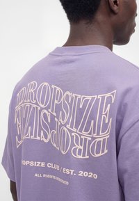 Personne portant un t-shirt lavande avec un grand texte déformé « DROPSIZE » et un texte plus petit « DROPSIZE CLUB / EST. 2020 TOUS DROITS RÉSERVÉS » dans le dos.