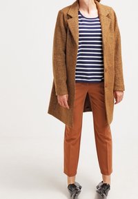 Brown wool blend coat with an oversized fit, paired with a navy and white striped shirt and brown tailored pants. Black shoes with bow accents.