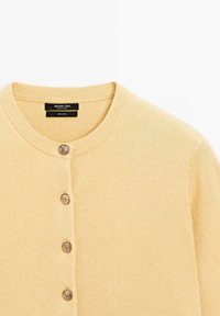 Ljusgul, 100% ull cardigan med rund hals, femknappslukning framtill och ribbade ärmslut och fåll. Mjuk textur, minimalistisk design.