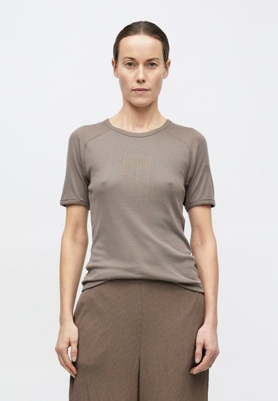 Sieviete ar atpakaļ piespraustu matu sakārtojumu, tērpusies taupe krāsas īsroku topā un atbilstošās biksēs, stāvot pret vienkāršu baltu fonu.