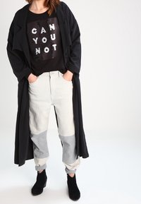 Abrigo largo negro sobre una camiseta negra con texto blanco. Pantalones en tonos gris claro y gris oscuro con bloque de color y puños elásticos. Botas negras hasta el tobillo.