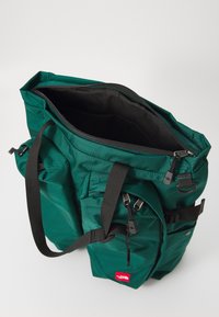Saco de pano verde com fecho em cima, apresentando alças ajustáveis, múltiplos bolsos e um logotipo visível na frente.