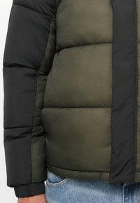 Zielono-czarna kurtka puchowa, rękaw i torso w komplecie z niebieskimi dżinsami, z widocznym pikowanym przeszyciem i elastycznym mankietem na nadgarstku.
