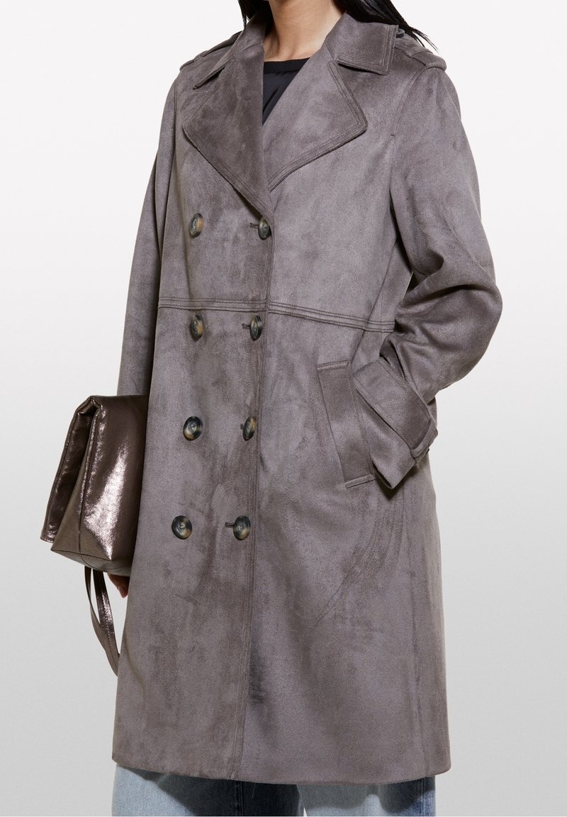 Femme portant un manteau en daim gris à double boutonnage avec col à revers et poches latérales, tenant un sac à main marron brillant, main dans la poche.