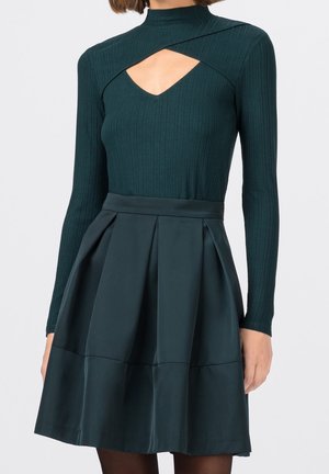 Femme portant une robe côtelée à manches longues vert foncé avec un décolleté découpé et une jupe plissée en ligne A.