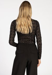 Vrouw met lang blond haar, gekleed in een zwart kanten top met lange mouwen en zwarte high-waisted broek, gezien vanaf de achterkant.
