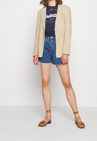 Blazer de lino beige sobre una camiseta gráfica azul marino, combinado con pantalones cortos de mezclilla de cintura alta y sandalias marrones de tiras.