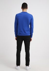 Sweat-shirt bleu à manches longues, col rond et poignets côtelés, associé à un jean noir slim et des baskets blanches. Vue de dos.