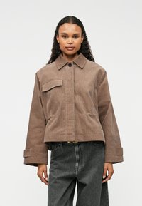 Chaqueta de pana marrón con cuello de camisa, bolsillo frontal y mangas anchas; combinada con pantalones de mezclilla grises. Textura suave y diseño estructurado.