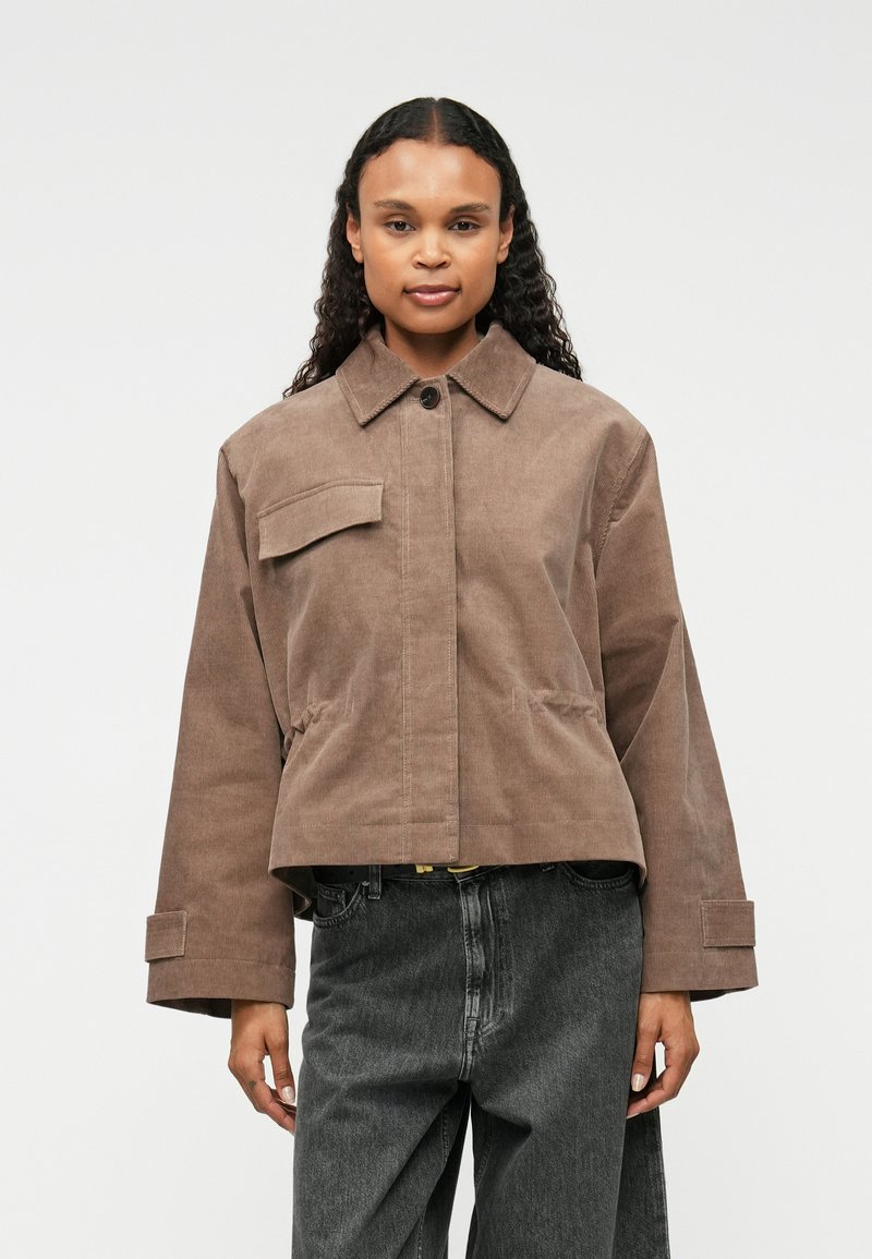 Chaqueta de pana marrón con cuello de camisa, bolsillo frontal y mangas anchas; combinada con pantalones de mezclilla grises. Textura suave y diseño estructurado.