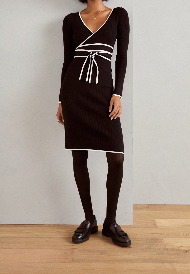 Robe noire côtelée à manches longues, col en V, accents blancs et taille nouée. Associée à des chaussures foncées et des collants noirs, sur un sol en bois.