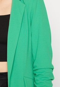 Blazer verde con tela lisa y ligeramente texturizada. Presenta un diseño entallado, solapas con muesca y un bolsillo lateral único. Anteriormente usado sobre una camiseta negra.