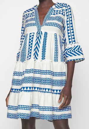 Vrouw draagt witte jurk met blauwe geometrische tribale patronen, V-hals en uitlopende 3/4-mouwen, staand tegen een witte achtergrond.