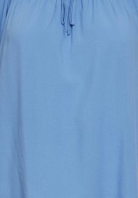 Blusa azzurra chiara con una texture morbida e liscia, scollatura arricciata con lacci e design fluido. Non presenta motivi distintivi o accenti.