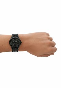 Schwarze analoge Armbanduhr an einer Hand, mit einem runden Ziffernblatt, strukturiertem schwarzen Armband, arabischen Ziffern und wasserdicht bis zu 5 ATM.
