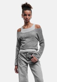 Grauer Strickpullover mit schulterfreiem Design und verschiedenen Texturen, kombiniert mit hellgrauen Jeans. Der Stoff wirkt weich und leicht.