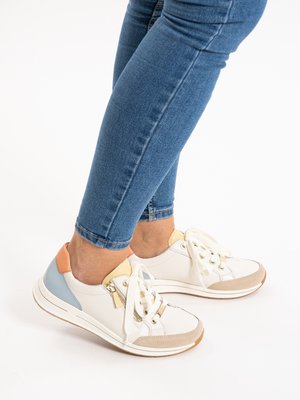 Persona con zapatillas blancas con detalles en beige, azul claro y melocotón, y jeans ajustados azules sobre un fondo blanco.