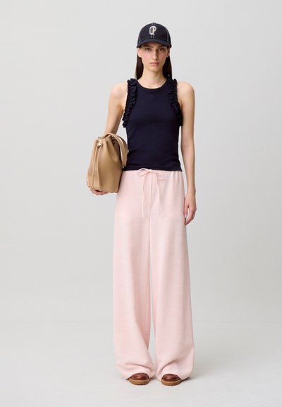 Femme portant un haut sans manches bleu marine avec des volants, un pantalon large rose, des chaussures marron, une casquette bleu marine et tenant un sac à main beige.