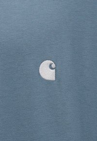 Λευκό κεντημένο λογότυπο Carhartt σε φόντο από μπλε πλεκτό ύφασμα, κεντραρισμένο και καθαρά ορατό.