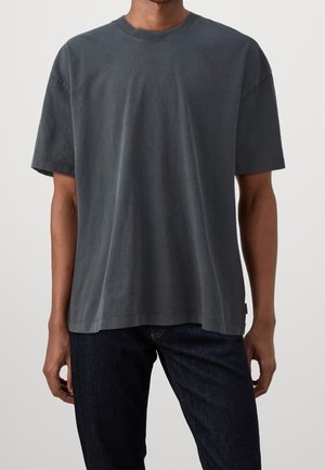 Mand iført en løs, kortærmet mørkegrå t-shirt og mørkeblå jeans mod en ensartet lys baggrund.