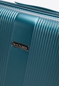 Valigia rigida color teal con texture rigata, dotata di una raffinata placca con logo nero. Chiusura con zip lungo il bordo superiore. Materiale durevole.
