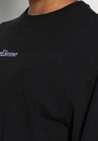 T-shirt noir en coton avec un col rond, orné d'un logo brodé violet sur la poitrine. Texture lisse, légère fluidité.