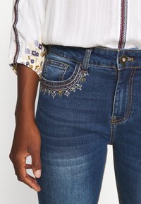 Tmavě modré džíny z denimu s propracovanou květinovou výšivkou podél okrajů kapes, s oranžovým prošíváním a klasickým střihem.