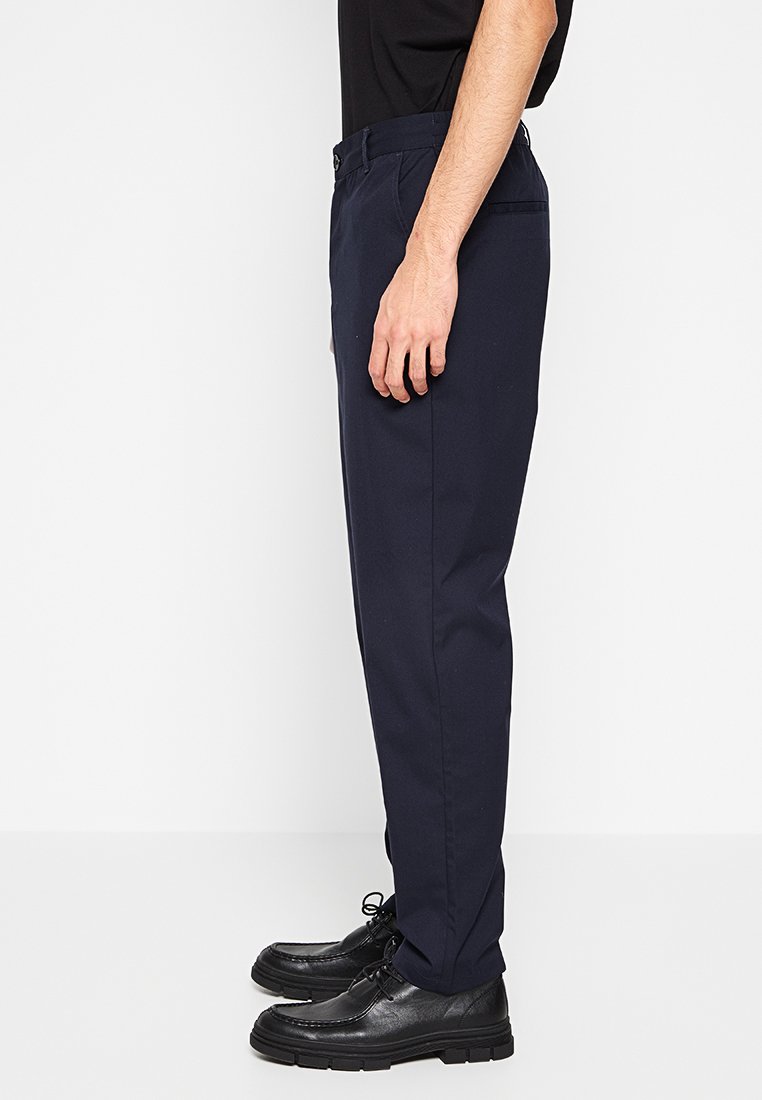 Dunkelblaue, strukturierte Hosen mit gerader Passform, versehen mit einem Knopfverschluss, Gürtelschlaufen und Seitentaschen. Getragen mit schwarzen Dressschuhen.
